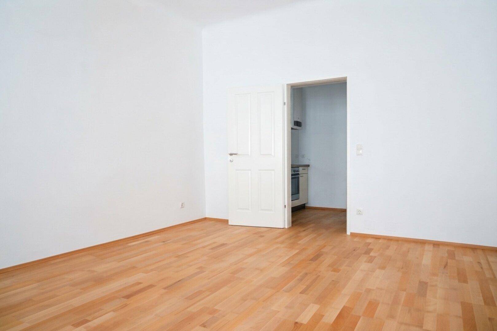 Geräumiges Zimmer mit Holzboden und Zugang zu einer kleinen Küchenzeile durch die offene Tür.