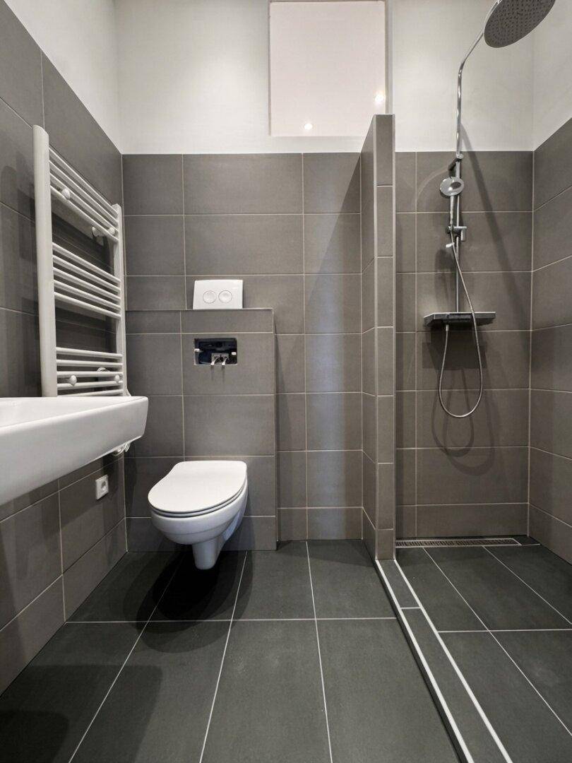 Modernes Badezimmer mit grauen Fliesen, wandhängendem WC, Waschbecken und begehbarer Dusche.
