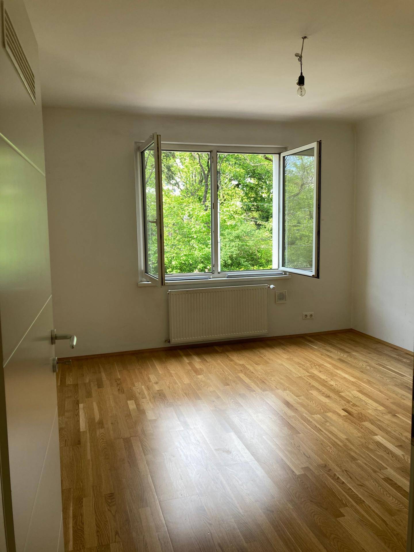 Helles Zimmer mit Parkettboden und Blick ins Grüne durch das geöffnete Fenster.