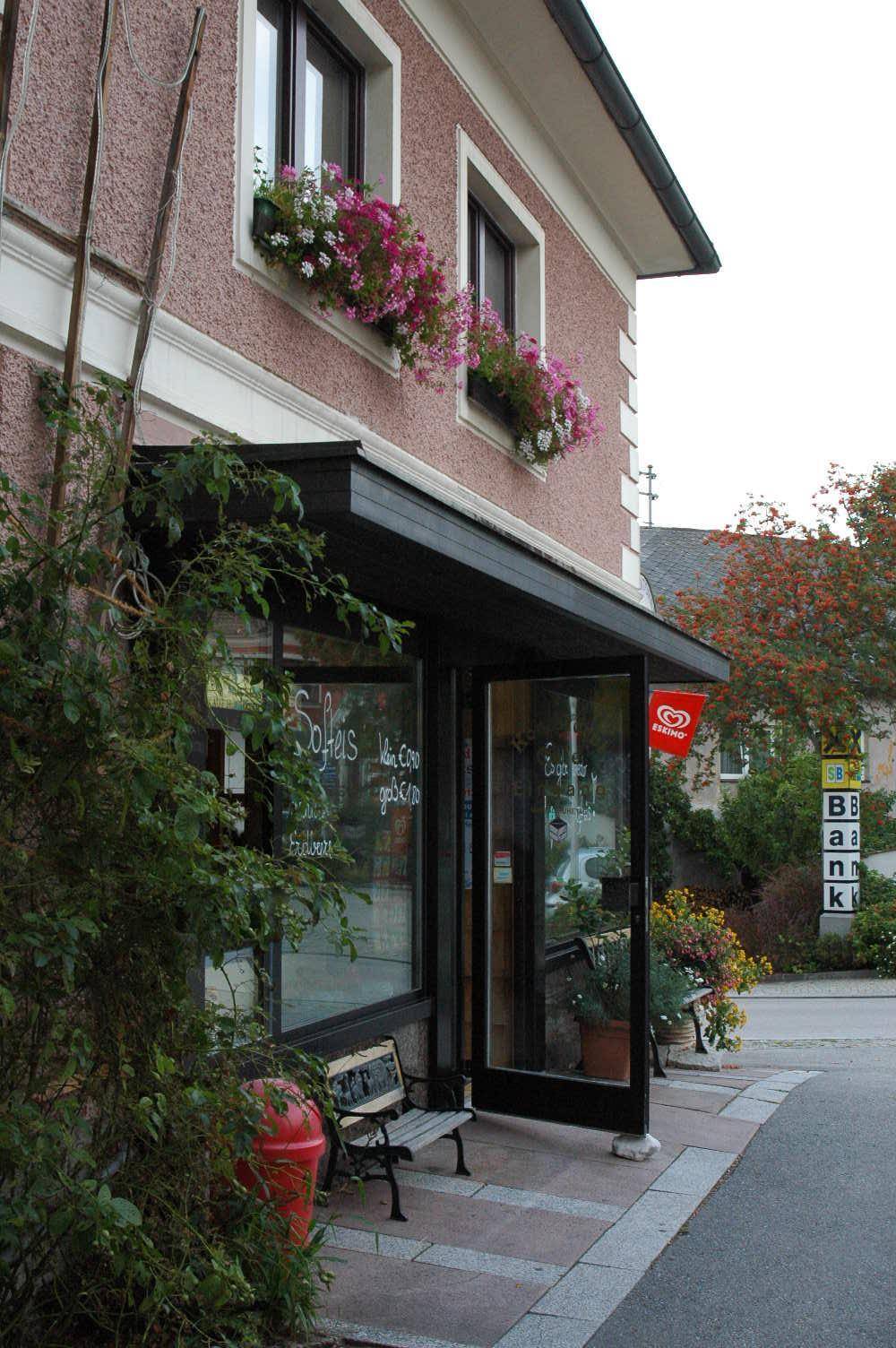 Nahaufnahme des Caféeingangs mit großen Schaufenstern, einer Sitzbank und Blumenkästen an der Fassade.