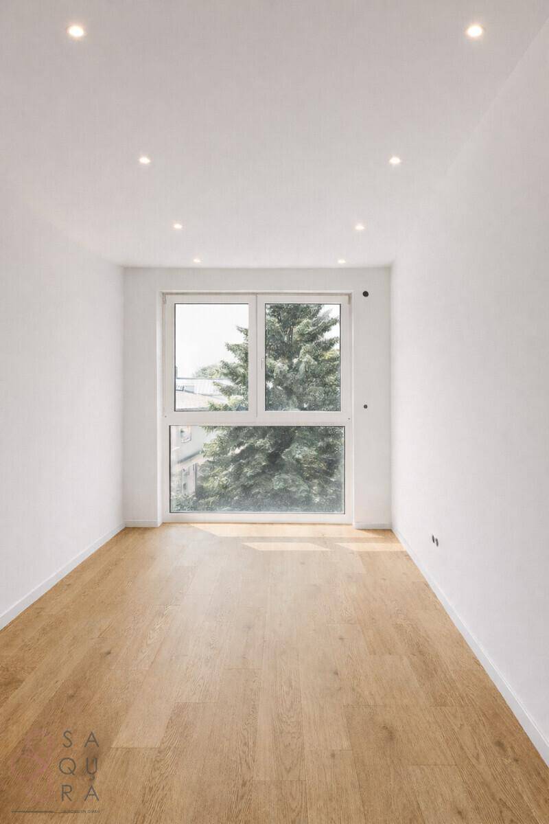 Leerer Raum mit Holzboden und großem Fenster, das viel Tageslicht hereinlässt und einen Baum zeigt.