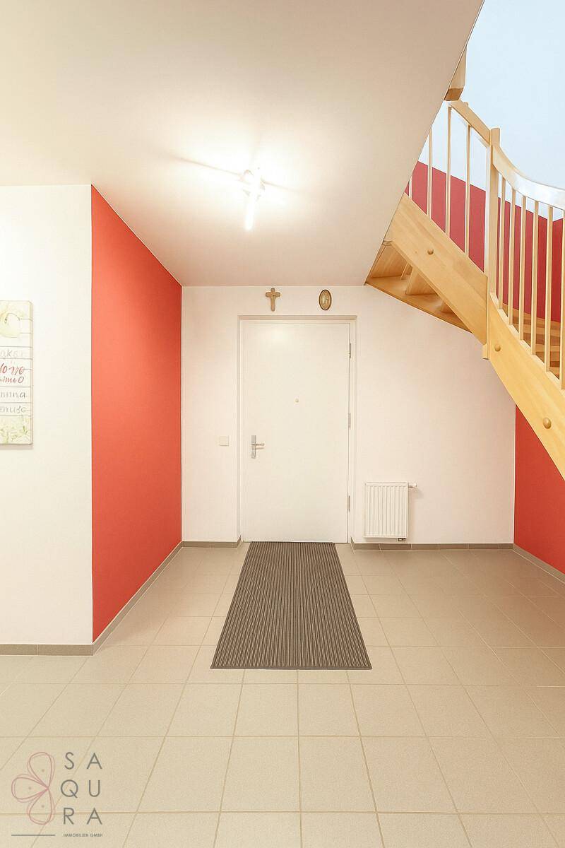 Eingangsbereich mit Fliesenboden, roter Akzentwand und Holztreppe zum Obergeschoss.