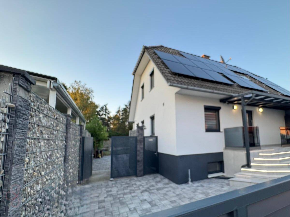 Seitenansicht eines modernen Hauses mit Photovoltaikanlage, Gabionenwand und gepflasterter Zufahrt.