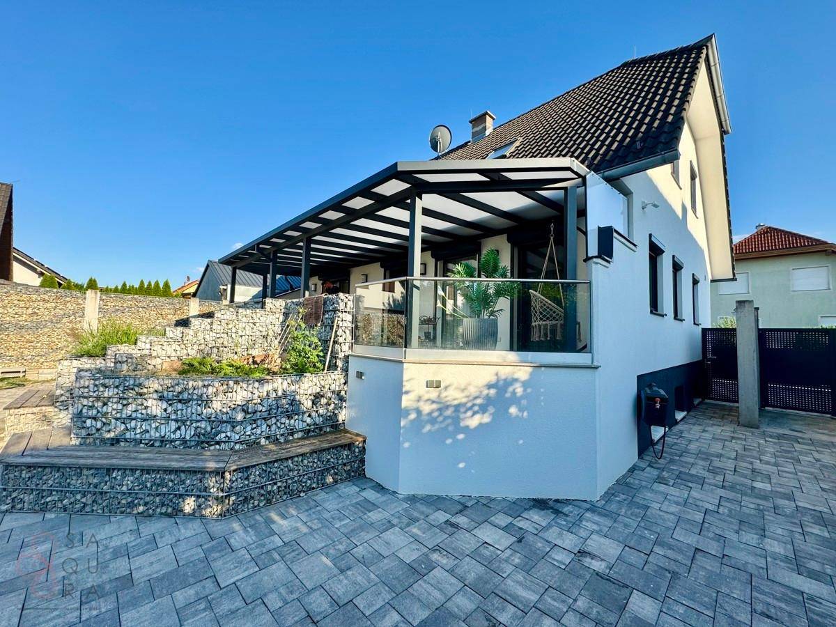 Moderne Hausfassade mit überdachter Terrasse, Gabionen-Landschaftsgestaltung und gepflastertem Zugang.