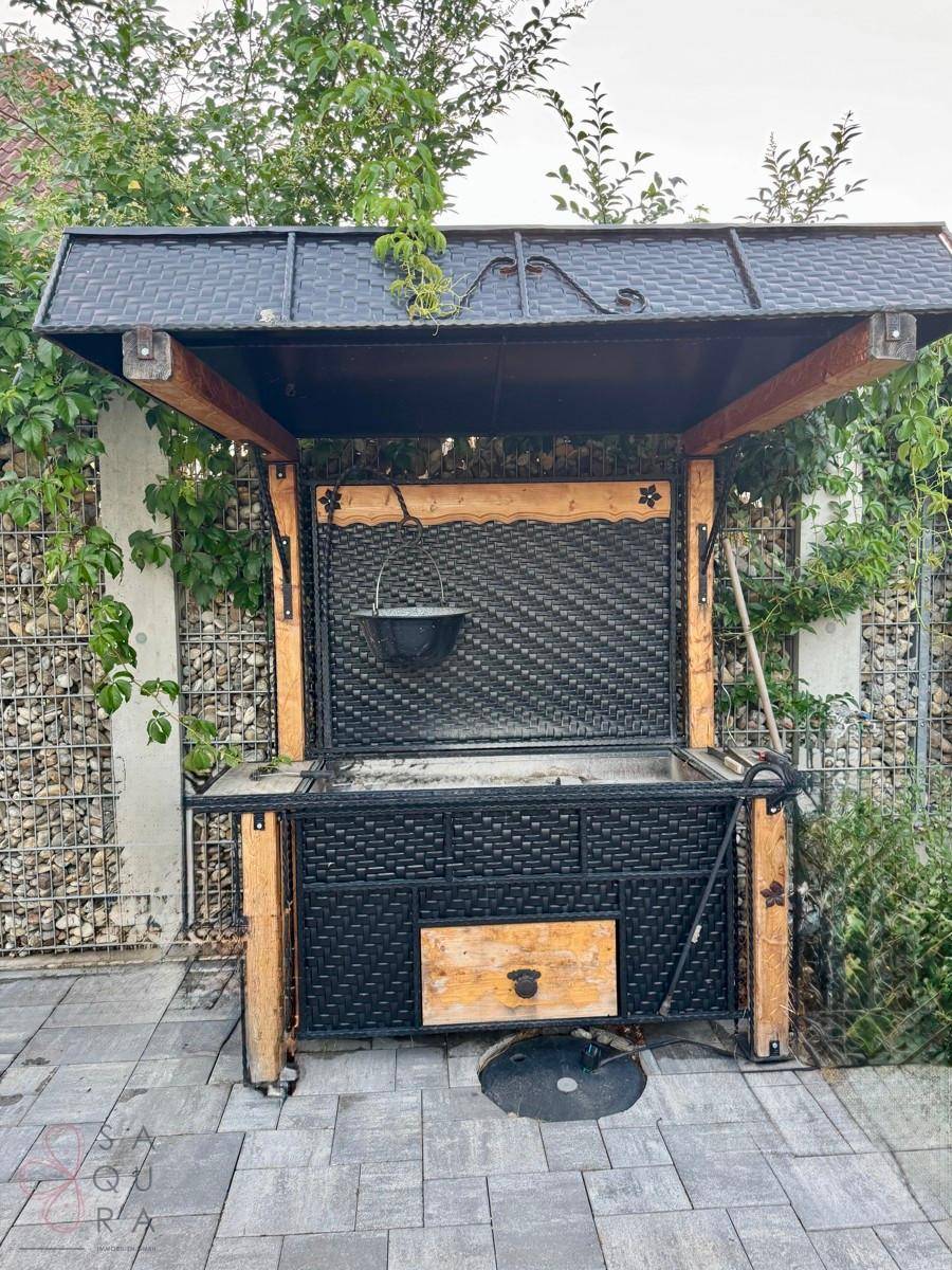 Rustikaler Grillplatz im Freien mit überdachter Konstruktion und Gabionenwand im Hintergrund.