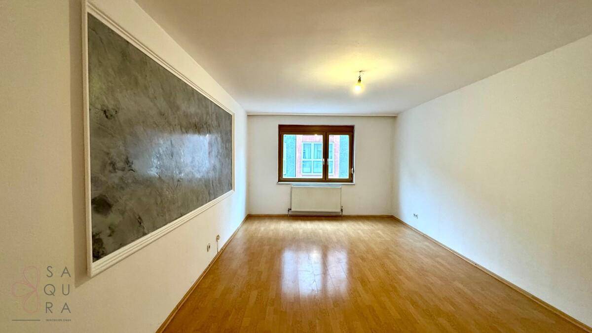 Leeres Wohnzimmer mit Holzboden, weißen Wänden und einem Fenster mit Blick nach draußen.