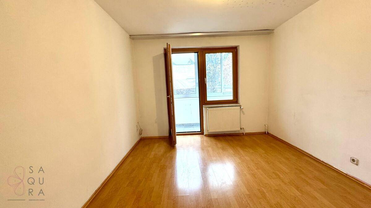 Leeres Schlafzimmer mit Holzboden, weißen Wänden und einer Tür, die zum Balkon führt.
