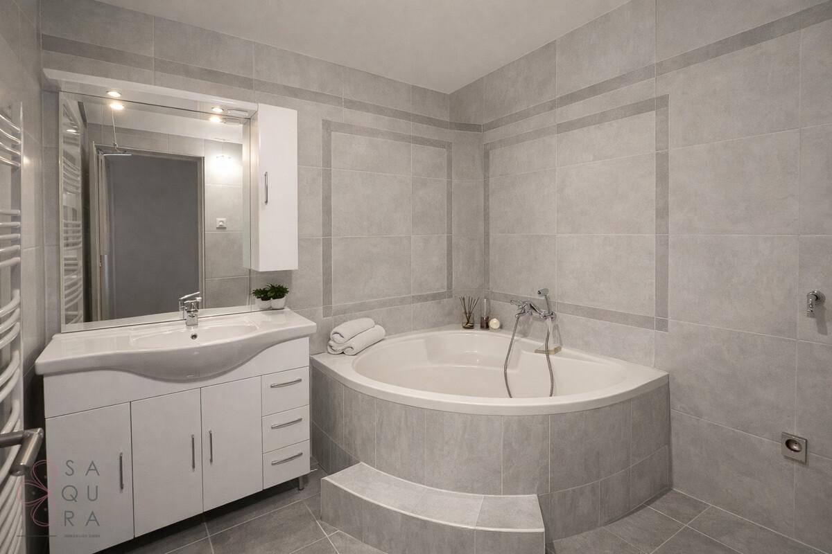 Modernes Badezimmer mit Eckbadewanne, grauen Fliesen und einem Waschtisch mit Spiegel.