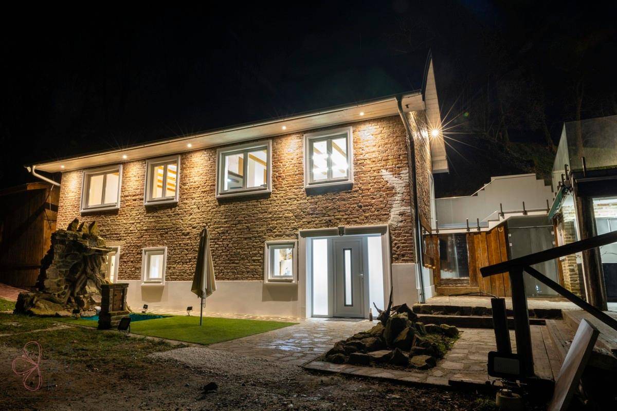 Beleuchtetes Backsteinhaus bei Nacht mit Wasserfall und gepflegtem Vorgarten.