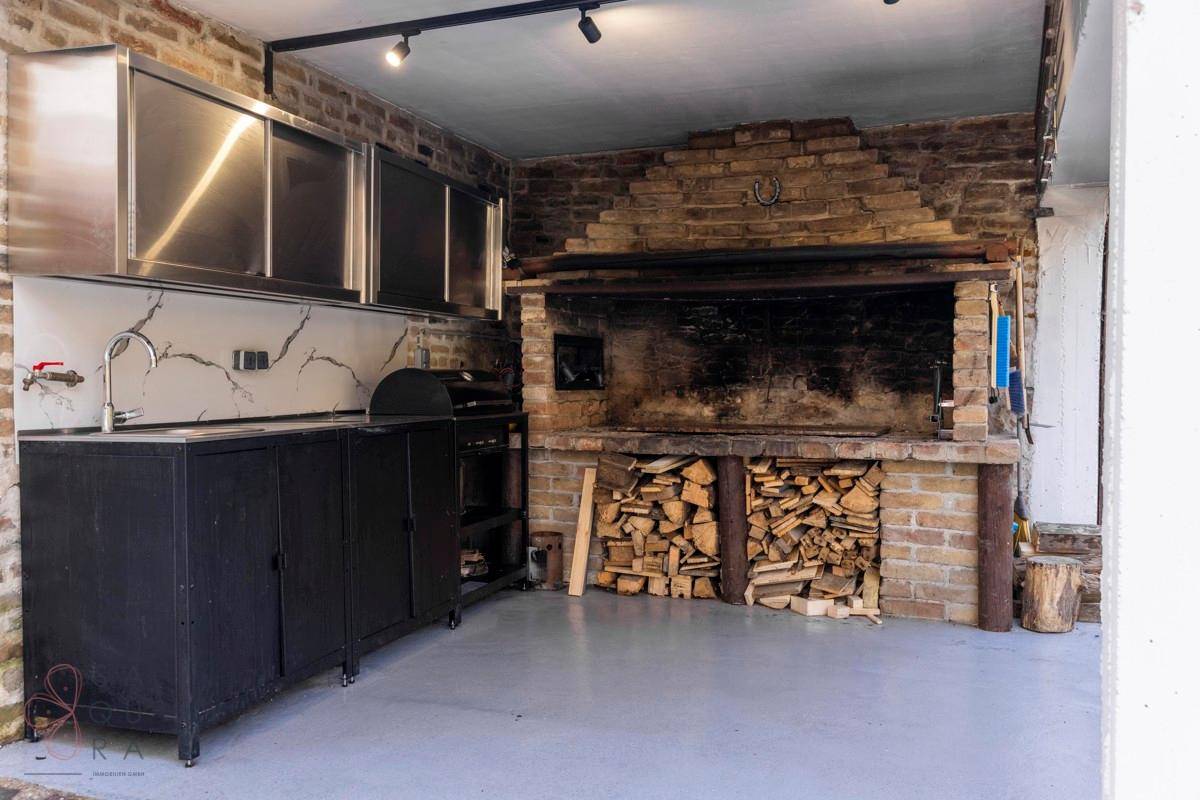 Rustikale Außenküche mit großem gemauertem Grill, Holzvorrat und modernen Edelstahlschränken.