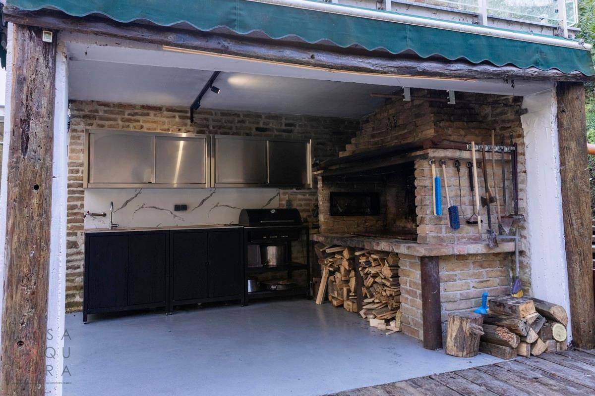 Geräumige Außenküche mit gemauertem Grill, Arbeitsbereich und Holzvorrat unter einer Überdachung.