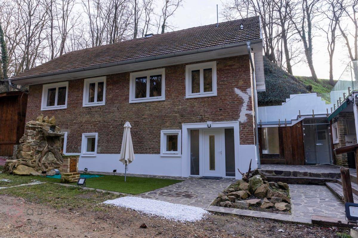 Backsteinhaus mit weißen Fenstern und Eingangstür, umgeben von Bäumen und einer kleinen Gartenanlage.