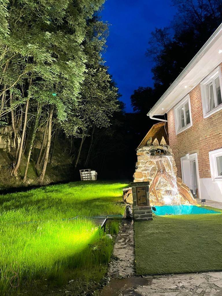 Seitlicher Gartenbereich des Hauses mit beleuchtetem Wasserfall und gepflegtem Rasen bei Nacht.