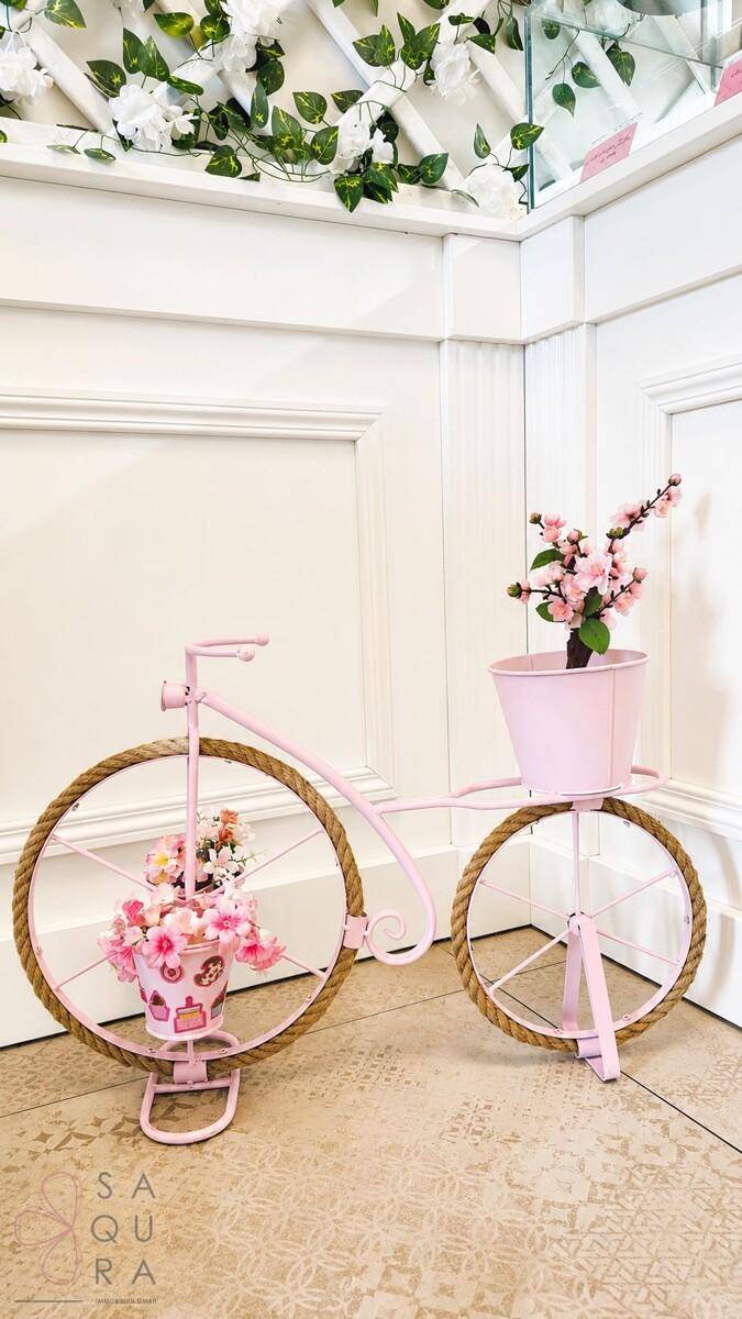 Dekorative Ecke im Verkaufsraum mit einer rosa Fahrradskulptur und Blumenarrangements auf Fliesenboden.