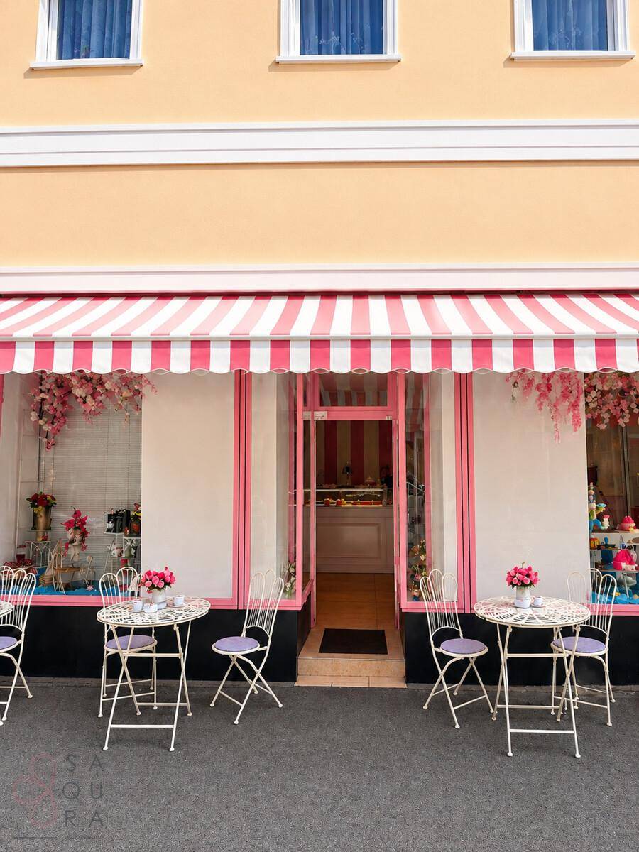Ansicht der Ladenfront mit rosa-weiß gestreifter Markise und mehreren Tischen für den Außenbereich.