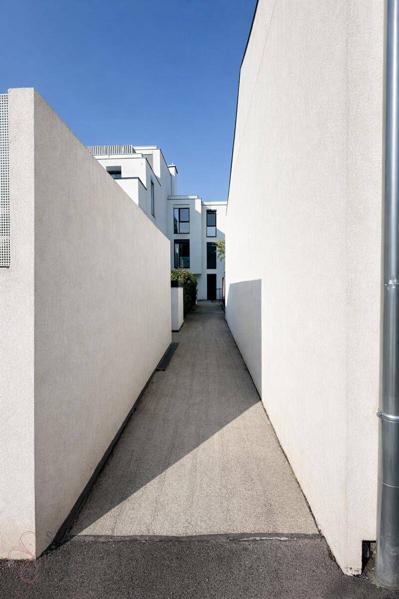 Schmaler Weg zwischen zwei modernen Gebäuden mit weißer Fassade unter blauem Himmel.
