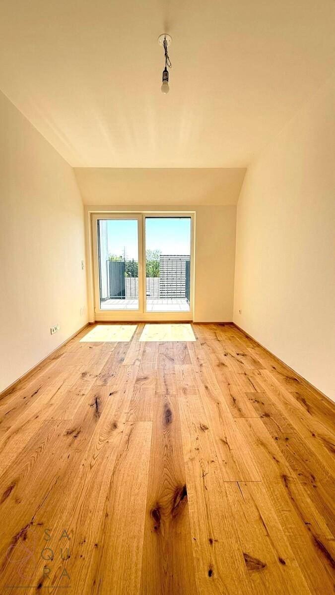 Leeres Schlafzimmer mit Holzboden und großen Fenstern, die viel Tageslicht hereinlassen.