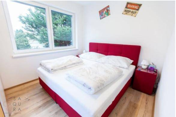 Schlafzimmer mit rotem Bett, Holzboden und Blick ins Grüne durch das Fenster.
