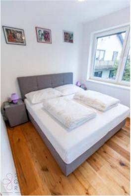 Schlafzimmer mit grauem Bett, Holzboden und großem Fenster.