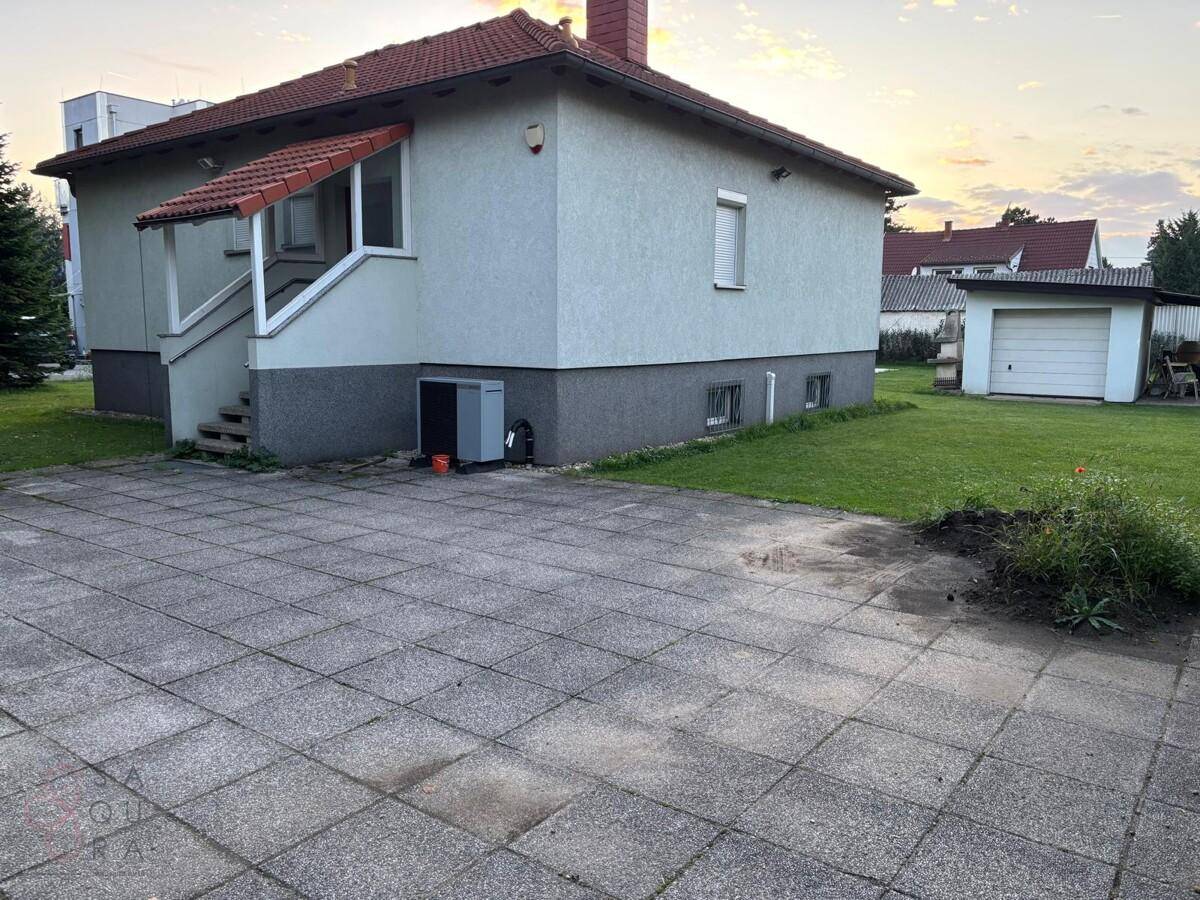 Rückansicht des Hauses mit gepflastertem Bereich, Rasenfläche und einem Nebengebäude im Abendlicht.