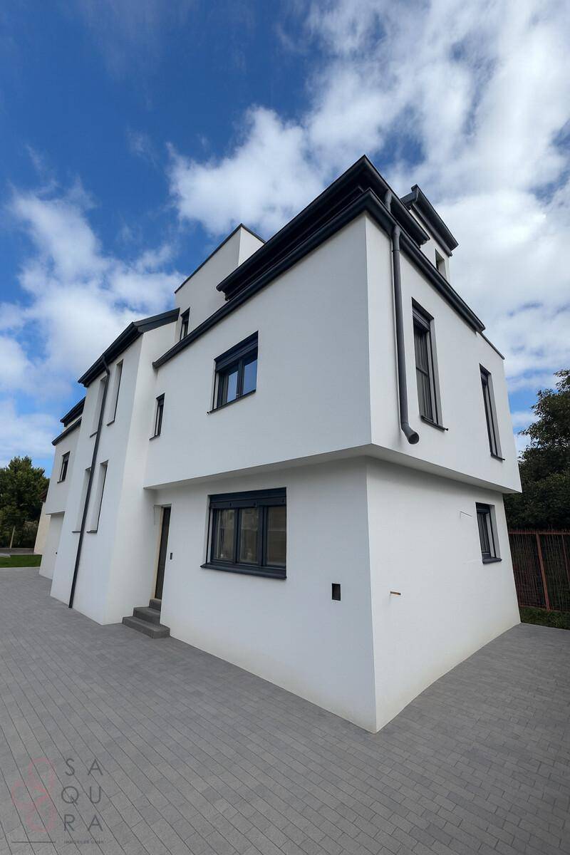 Eine seitliche Ansicht des modernen Gebäudes mit weißer Fassade und gepflastertem Vorplatz.