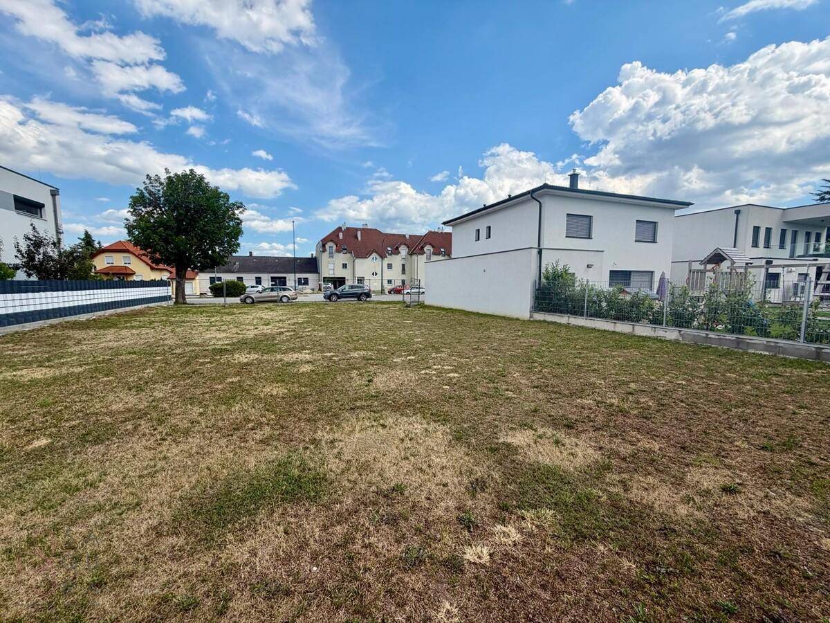 Weitläufiges unbebautes Grundstück mit trockenem Gras, umgeben von modernen Häusern unter blauem Himmel.