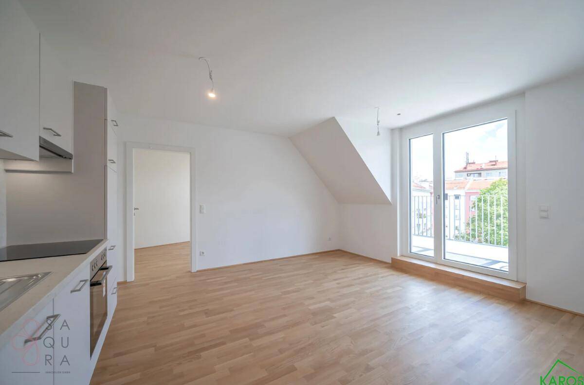 Heller, leerer Wohn-Essbereich mit offener Küchenzeile, Holzboden und Zugang zum Balkon.