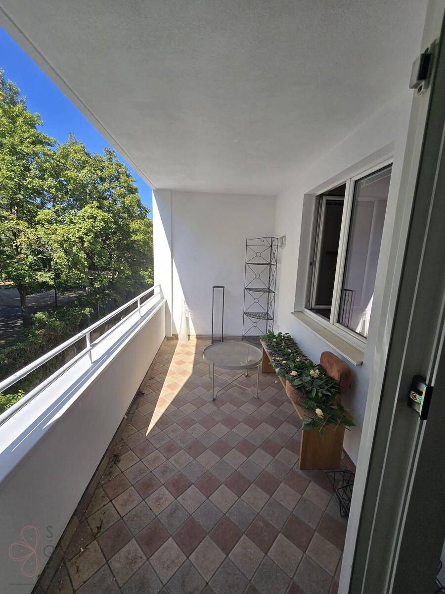Sonniger Balkon mit Fliesenboden, kleinem Tisch und Blick ins Grüne mit Bäumen.