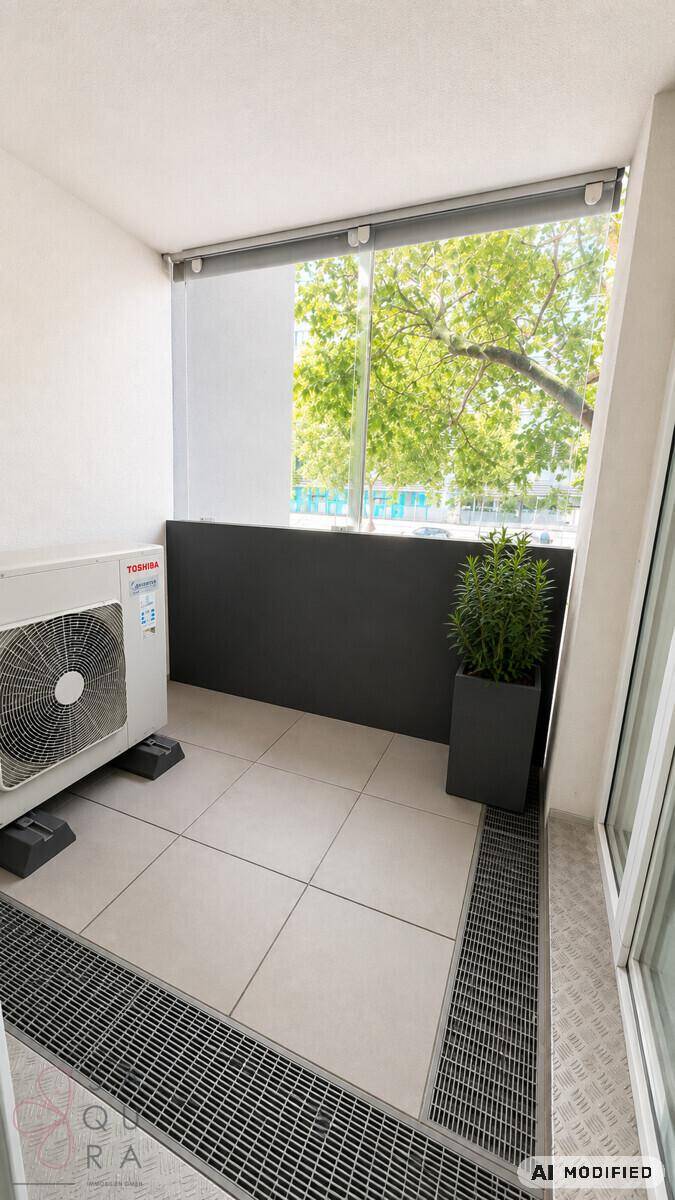 Kleiner Balkon mit Klimaanlage, Fliesenboden und Blick auf grüne Bäume.