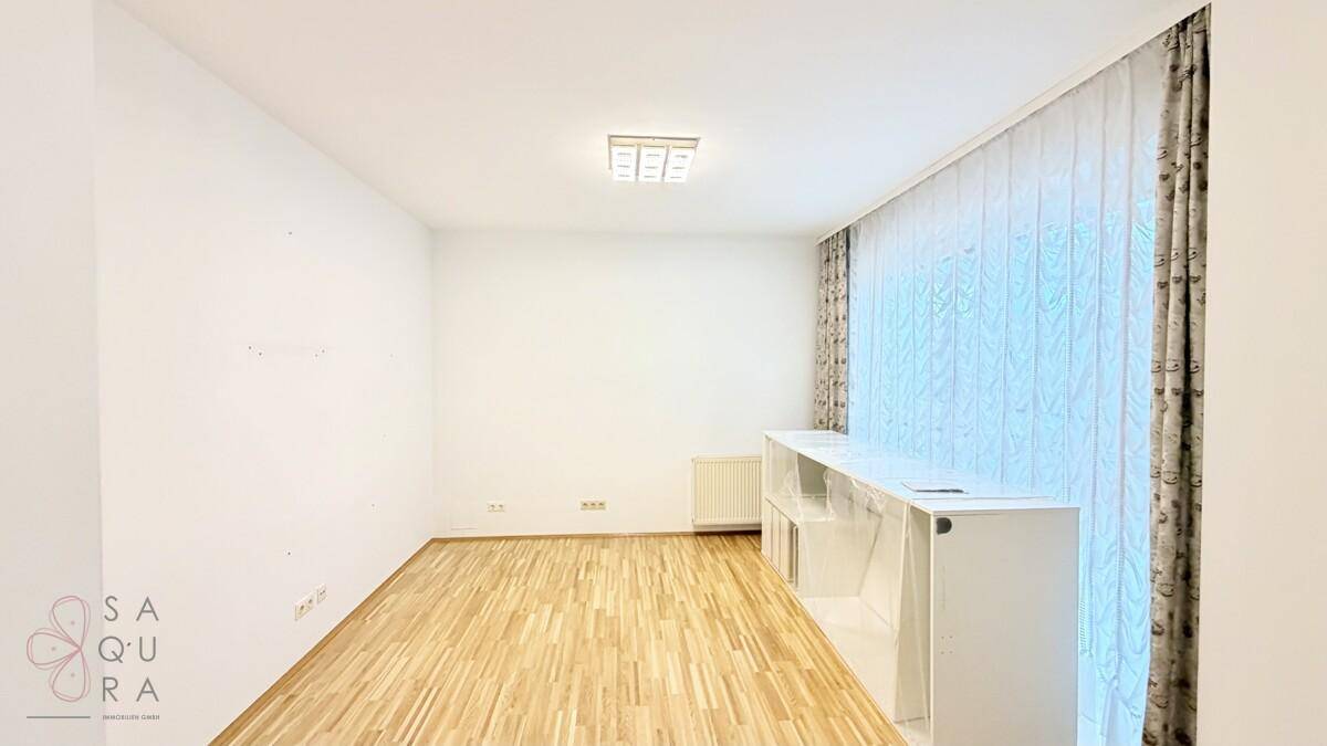 Leerer Raum mit Holzboden, weißen Wänden, Heizkörper und einem Fenster mit Vorhängen.