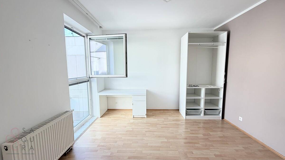 Helles Schlafzimmer mit Holzboden, Fenster, Heizkörper und offenem Kleiderschrank.
