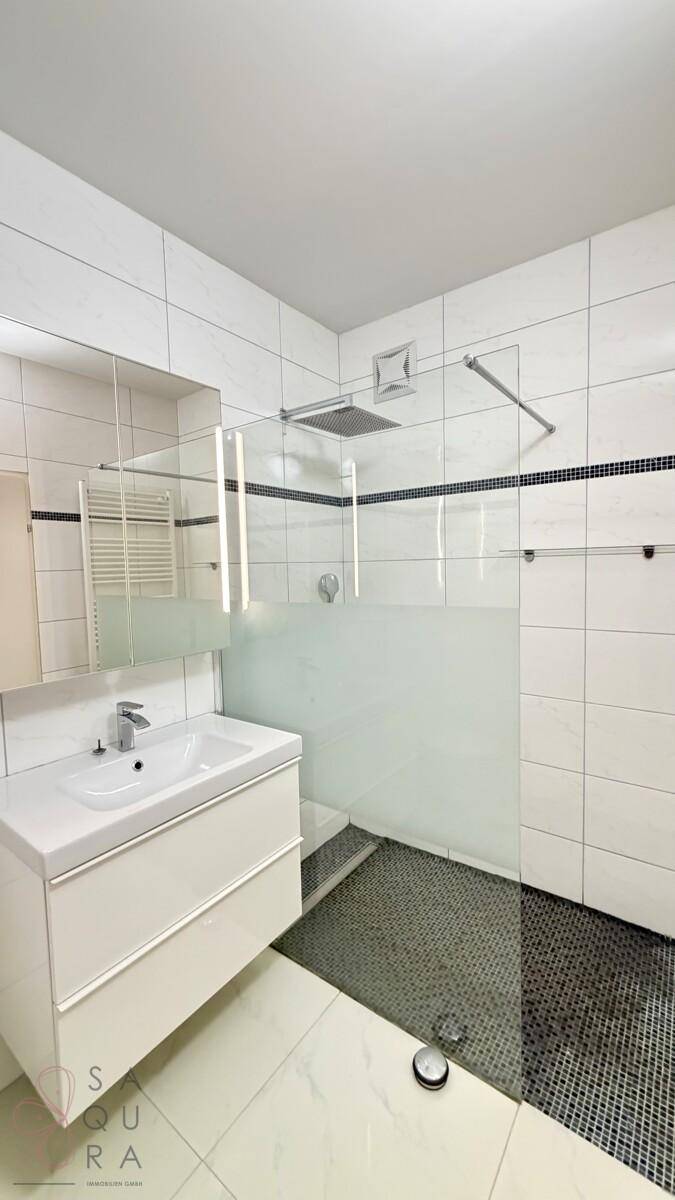 Modernes Badezimmer mit ebenerdiger Dusche, Waschtisch und Spiegelschrank, hell gefliest.