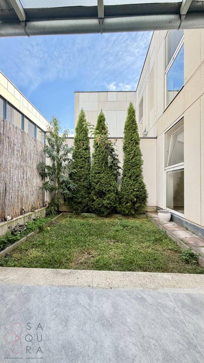 Blick vom überdachten Bereich auf den kleinen Garten mit Rasen und hohen Bäumen.