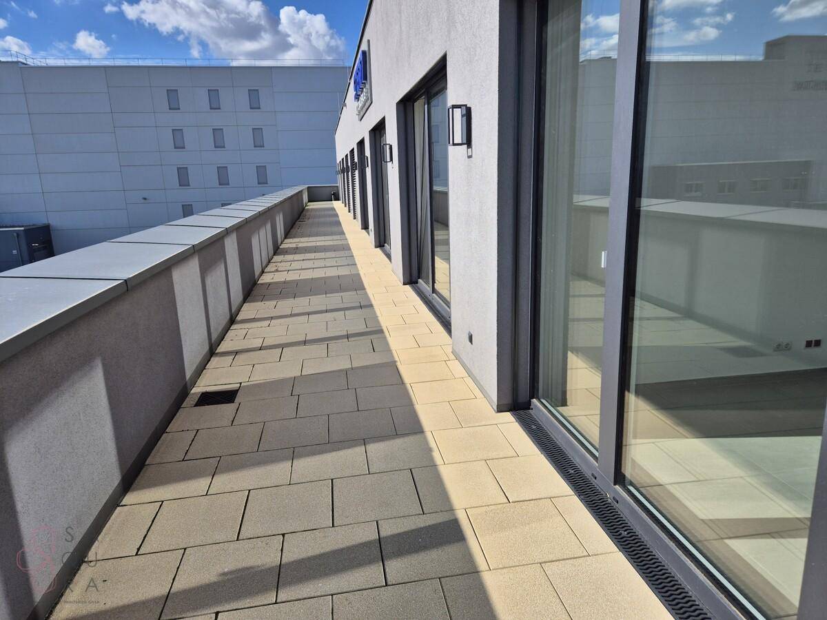 Lange, gepflasterte Terrasse entlang der modernen Gebäudefassade mit Blick auf die Umgebung.