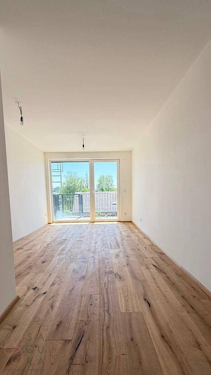 Leerer Raum mit Holzboden und großen Fenstern, die Zugang zu einem Balkon bieten.