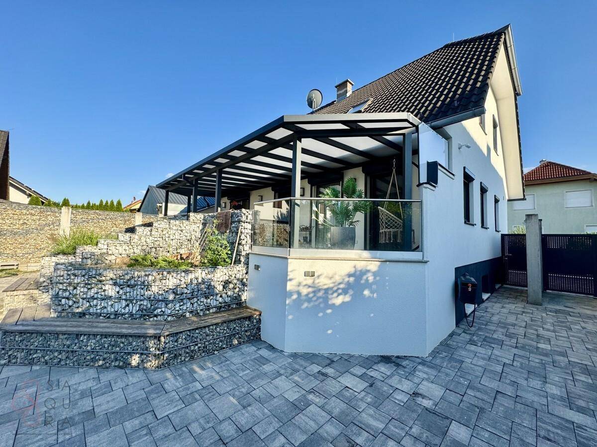 Moderne Hausfassade mit überdachter Terrasse, Glasgeländer und Gabionenmauern im Außenbereich.