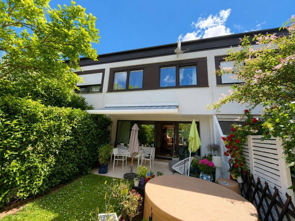 Hausfassade mit großer Terrasse, Whirlpool und üppiger Gartenbepflanzung.