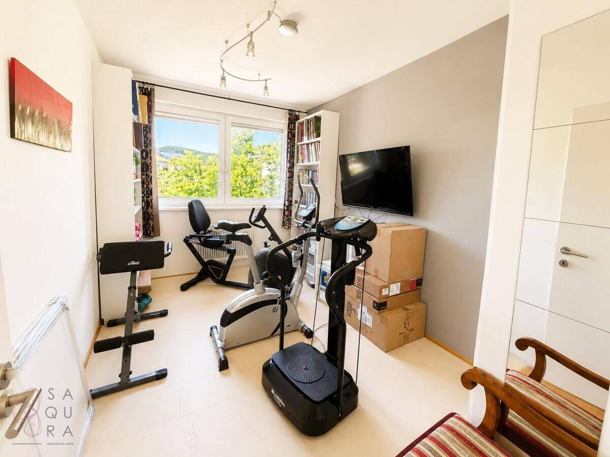 Heller Raum mit Fitnessgeräten, Bücherregalen und einem Fenster mit Blick ins Grüne.
