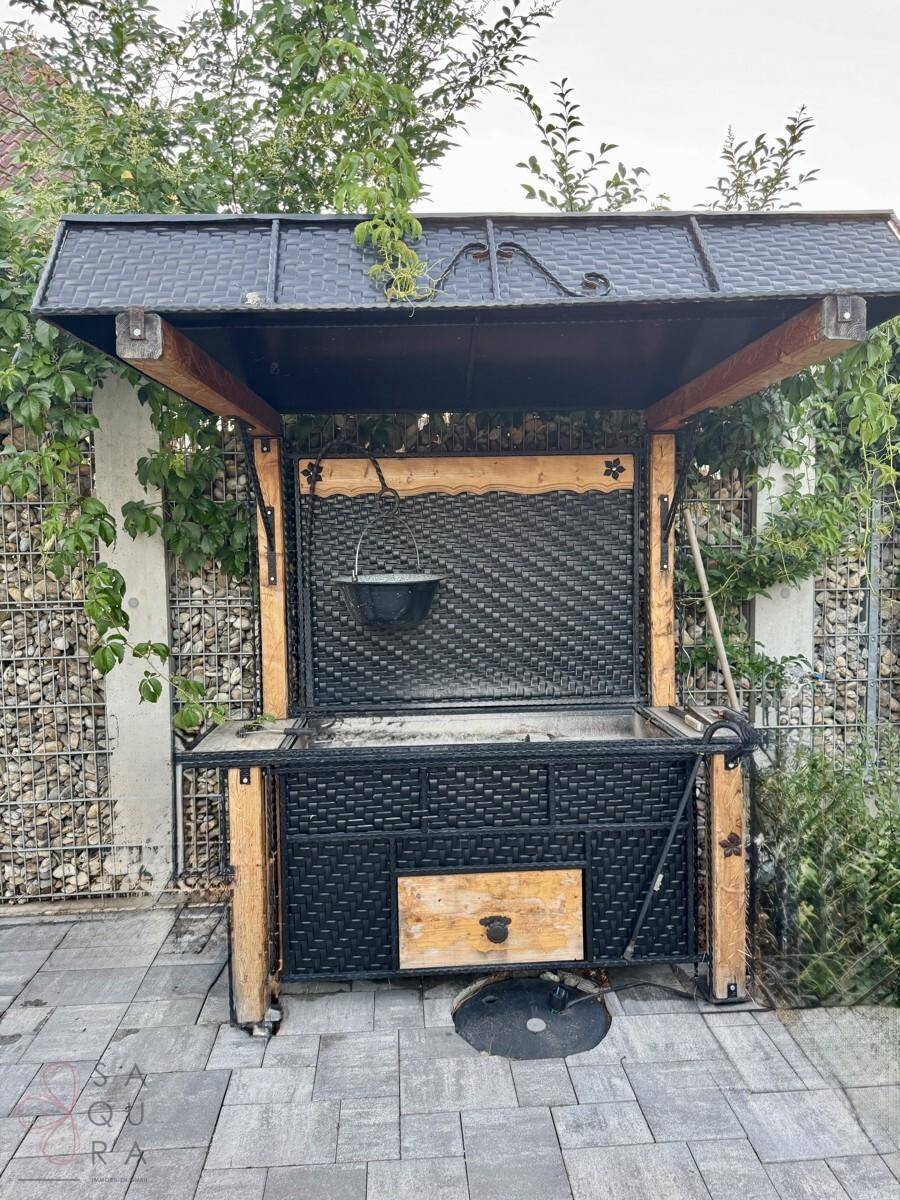 Gemütlicher Grillbereich im Freien mit überdachtem Grill und Gabionenmauer im Hintergrund.