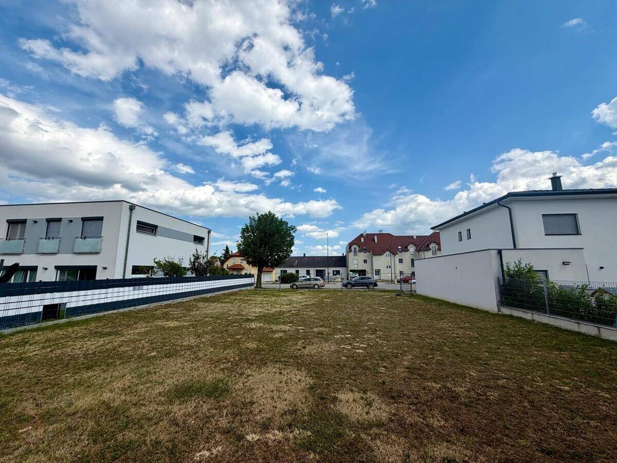 Offenes Baugrundstück mit trockenem Gras und einer Reihe von Wohnhäusern unter einem bewölkten Sommerhimmel.