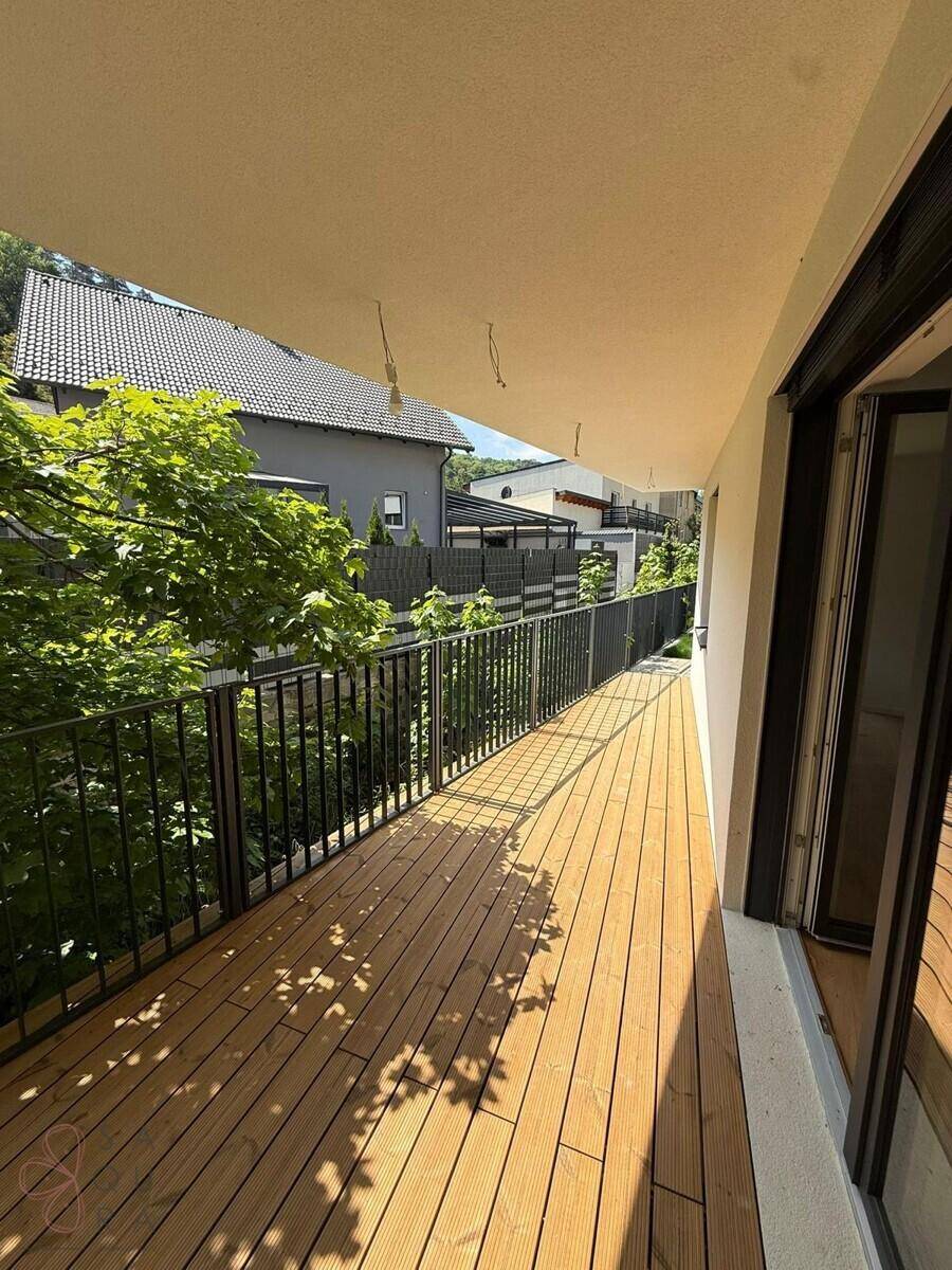 Langer, sonniger Balkon mit Holzboden und schwarzem Geländer, umgeben von viel Grün.