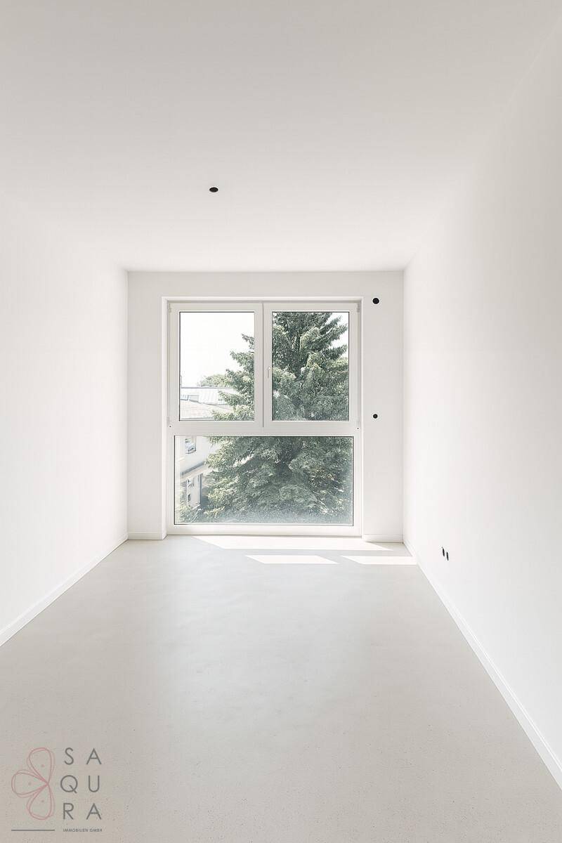 Leerer Raum mit hellem Boden und großem Fenster, das einen Baum und Tageslicht hereinlässt.