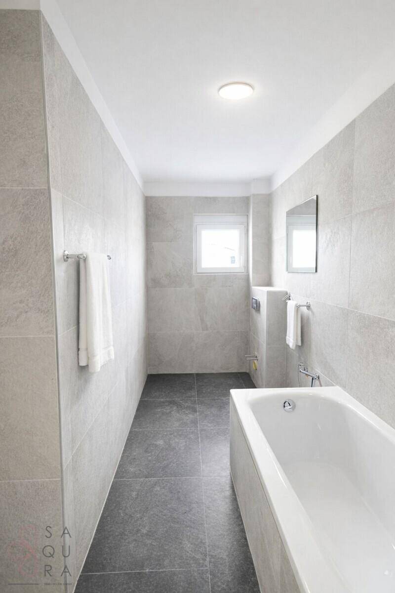 Modernes Badezimmer mit grauen Fliesen, Badewanne und Fenster für natürliches Tageslicht.