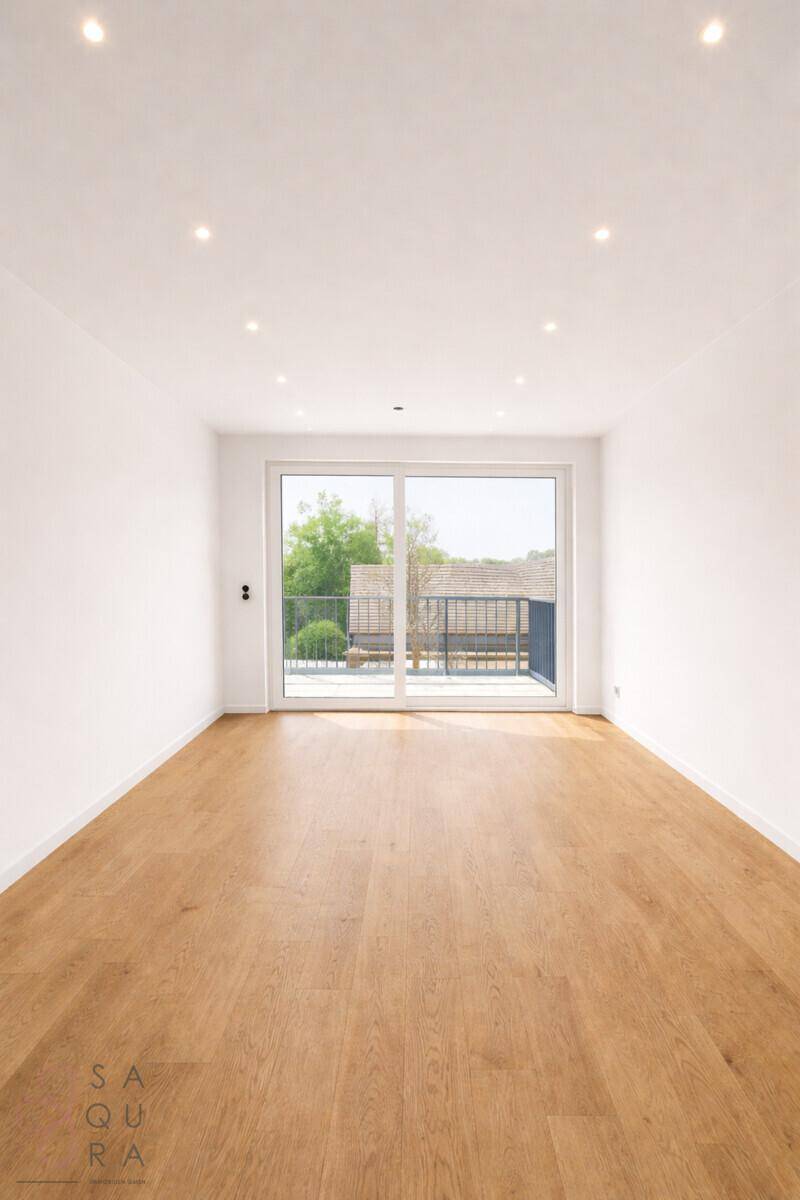 Helles, leeres Zimmer mit Holzboden und großen Fenstern, die auf einen Balkon führen.