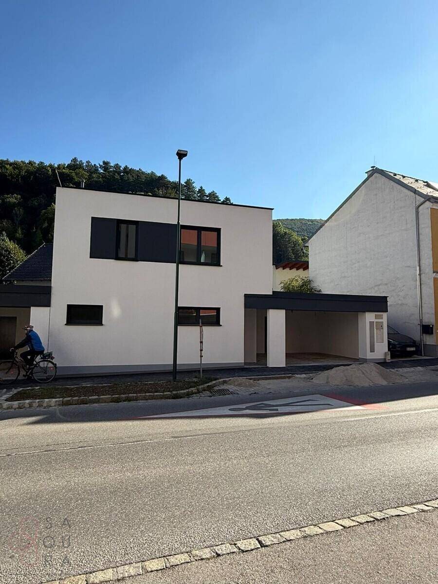 Einzelnes modernes Haus mit Carport an einer Straße, umgeben von älteren Gebäuden und Natur.