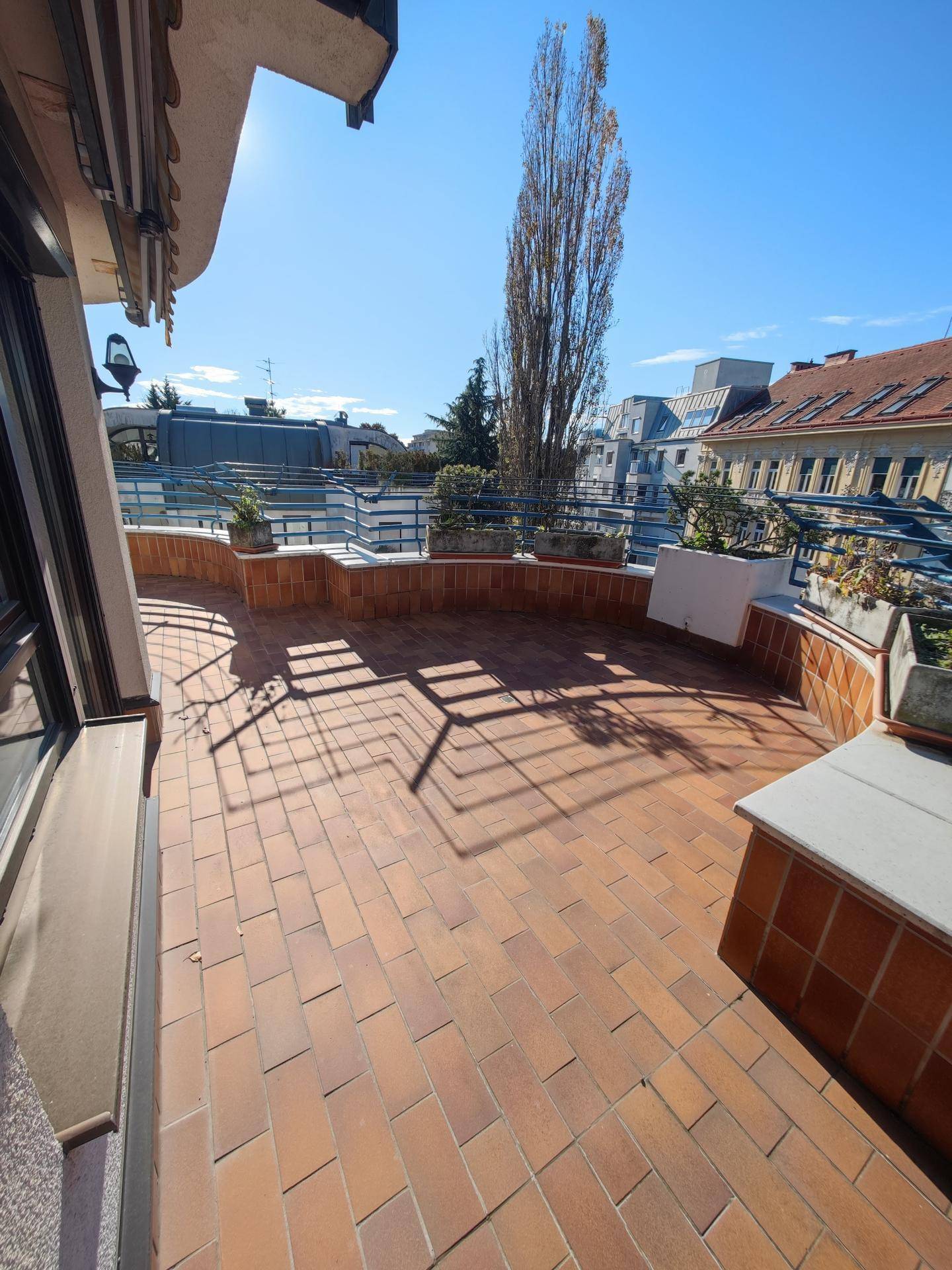 Weitläufige Dachterrasse mit Ziegelboden und Blick auf die städtische Umgebung unter blauem Himmel.