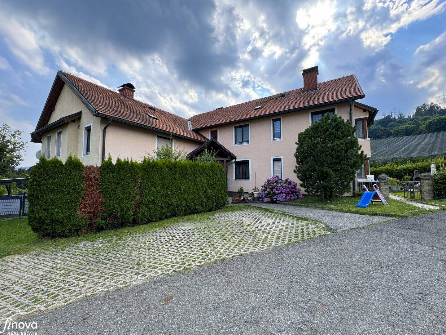 Hausfassade mit gepflasterter Auffahrt, Hecke und Gartenbereich unter bewölktem Himmel.