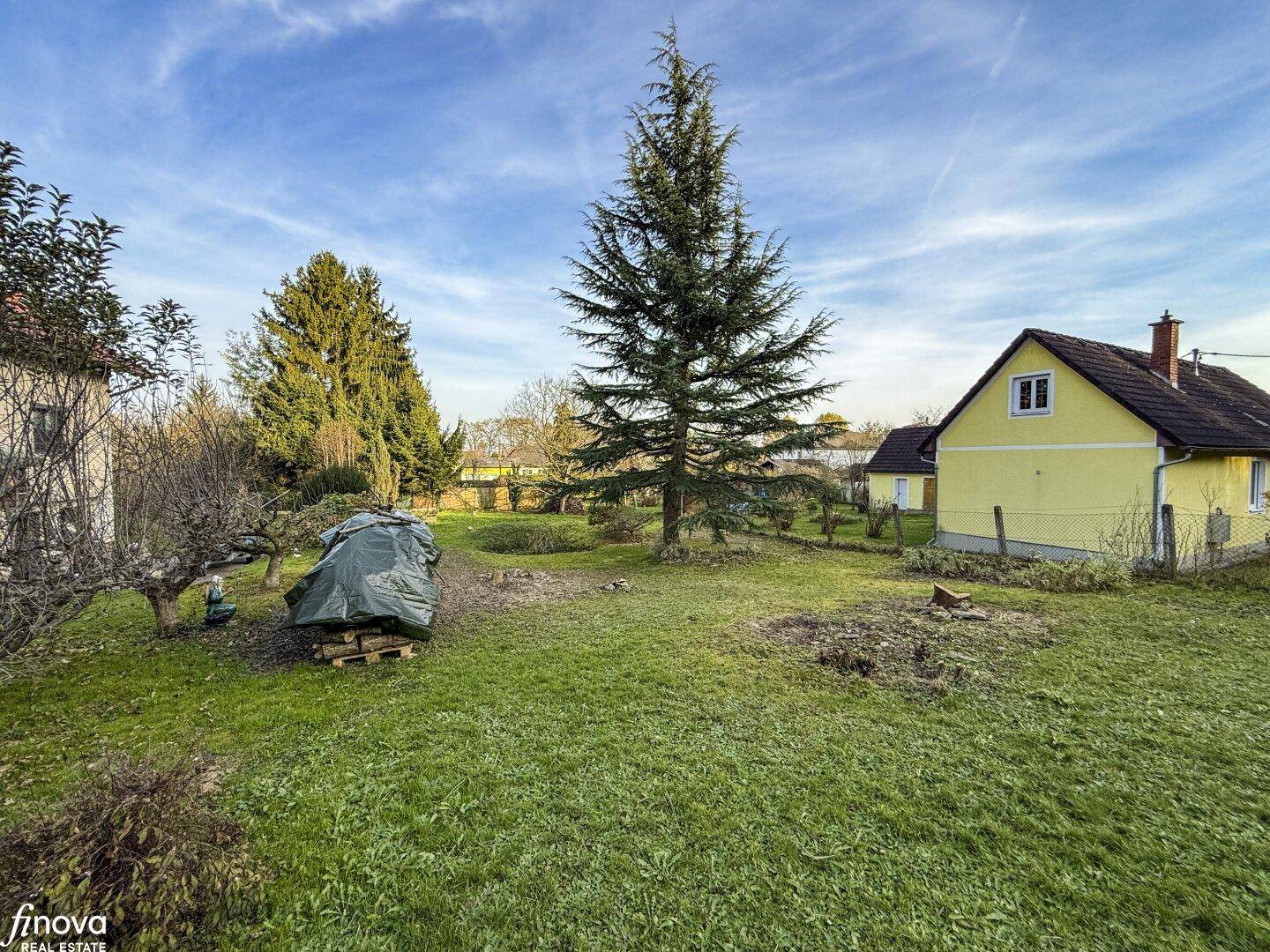 Weitläufiger Garten mit altem Baumbestand und einem traditionellen gelben Haus unter klarem Himmel.