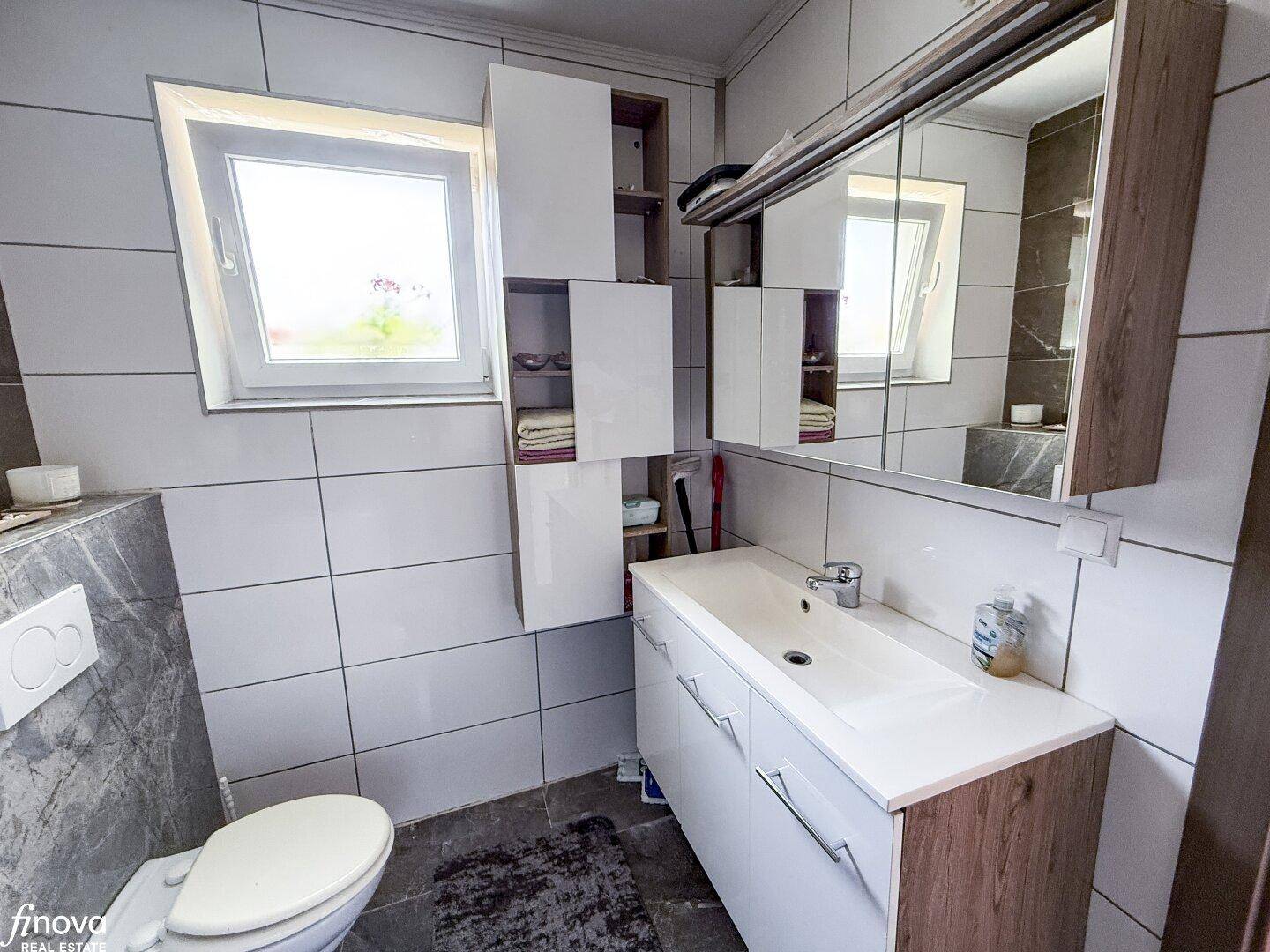 Helles Badezimmer mit Waschbecken, Spiegelschrank, Toilette und Fenster.