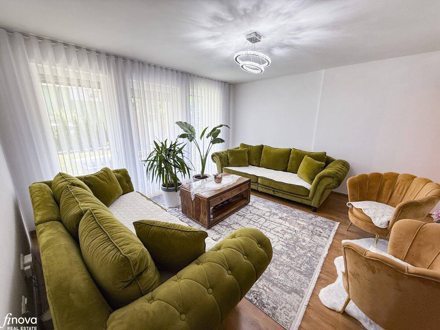 Helles Wohnzimmer mit großen Fenstern, grünen Sofas und modernem Teppich.